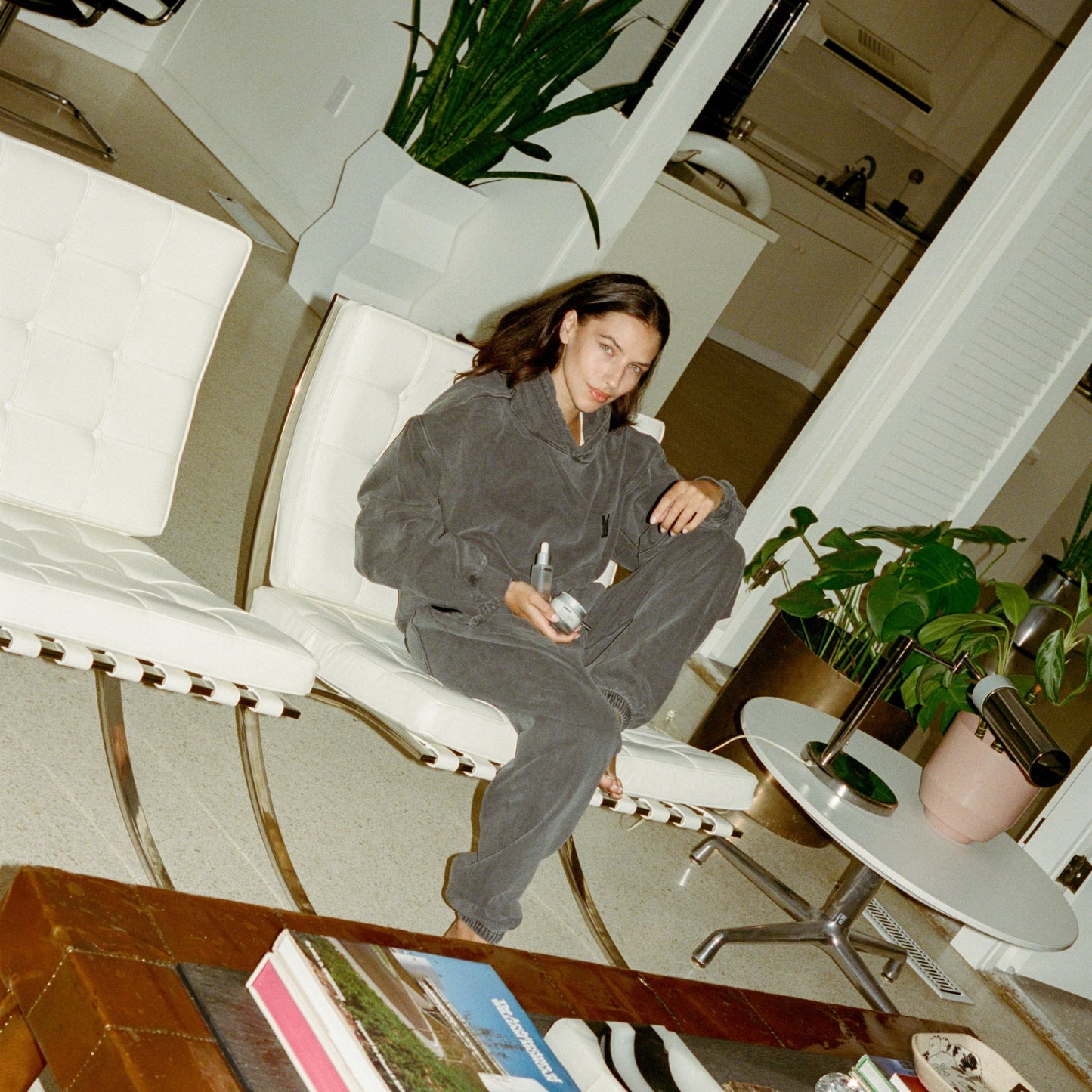 Person sitting on a white chair wearing comune gear in a room with plants and furniture, holding 'COMUNE' Essentials Skincare products.