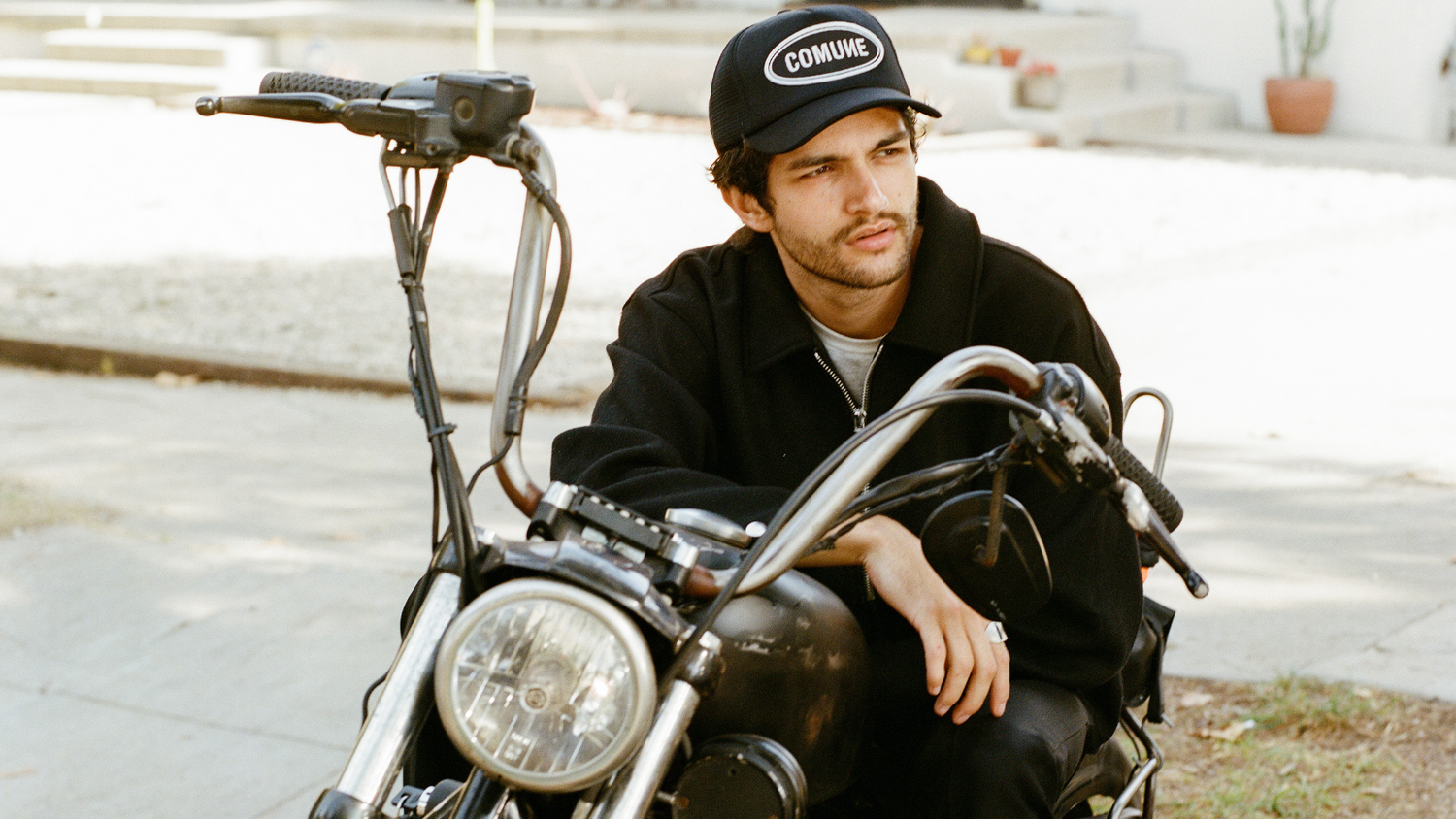 COMUNE Artist Brandon Shoop sitting on a motorcycle wearing a cap with 'COMUNE' branding.