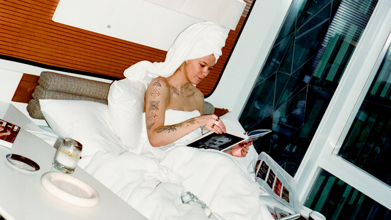 Comune model in a white robe sitting on a bed reading a magazine with a modern room setting.