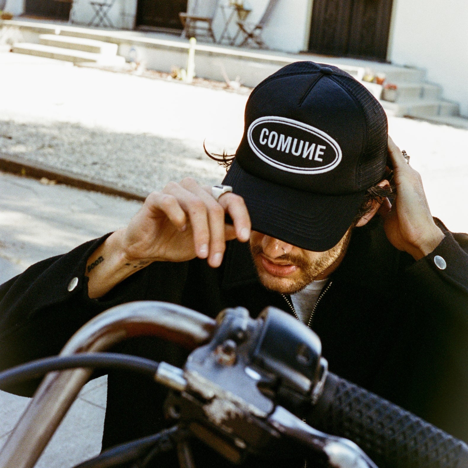 Person wearing the COMUNE trucker hat sitting on his custom motorcycle, outdoors.