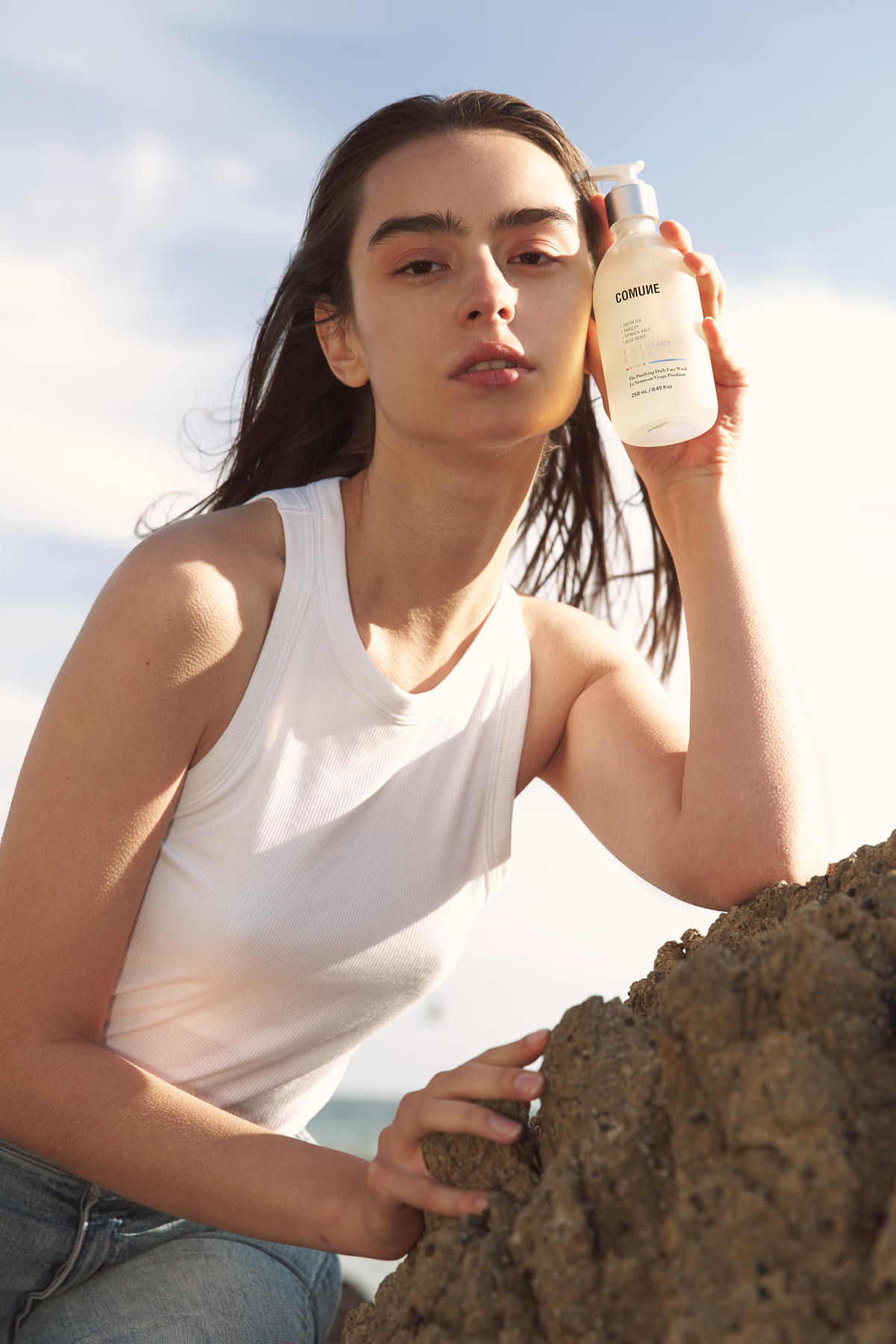 Comune Essentials Face Cleanser held by a women on top of rock near the ocean