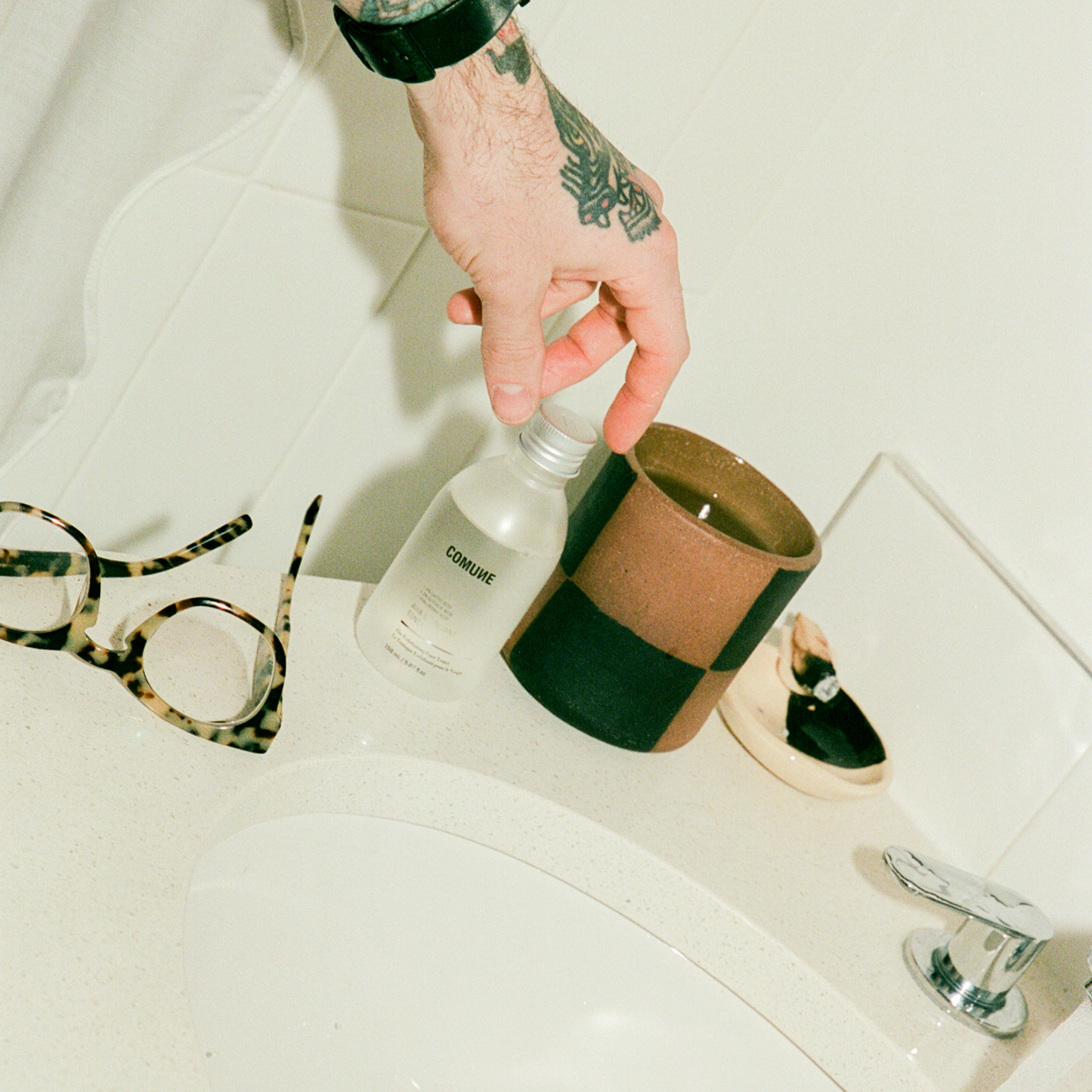 Hand holding a bottle labeled 'COMUNE AHA Exfoliant Toner' on a bathroom counter with glasses and candle holder in the background.