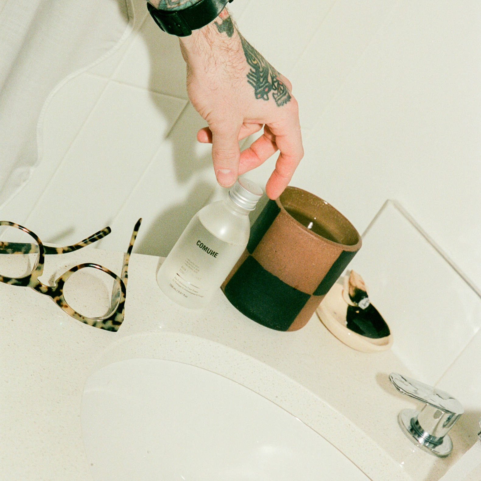 Hand holding a bottle labeled 'COMUNE AHA Exfoliant Toner' on a bathroom counter with glasses and candle holder in the background.
