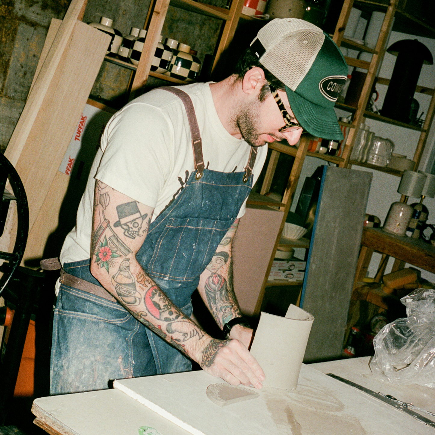 Man with tattoos wearing overalls and a cap in a workshop setting