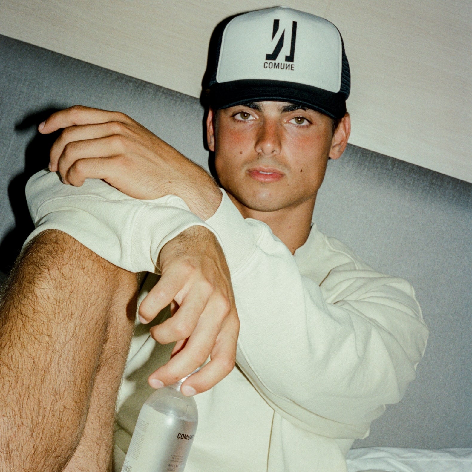 Person wearing a COMUNE cap and holding Comune AHA Exfoliant Toner with a visible brand logo, sitting on a couch.