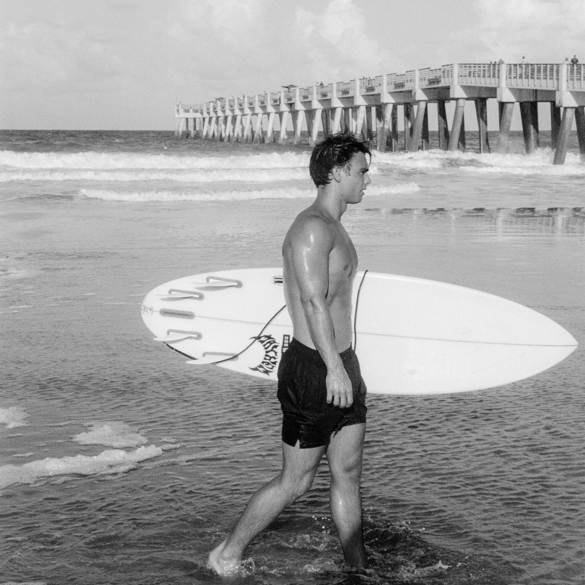 Man walking on a beach with a surfboard, with a pier in the background.