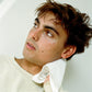 Man with wavy hair wearing a white shirt against a light background, drying his face after using Comune Essentials Face Cleanser