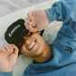 Person wearing a black Comune bucket hat with 'Comme' text, smiling at the beach.