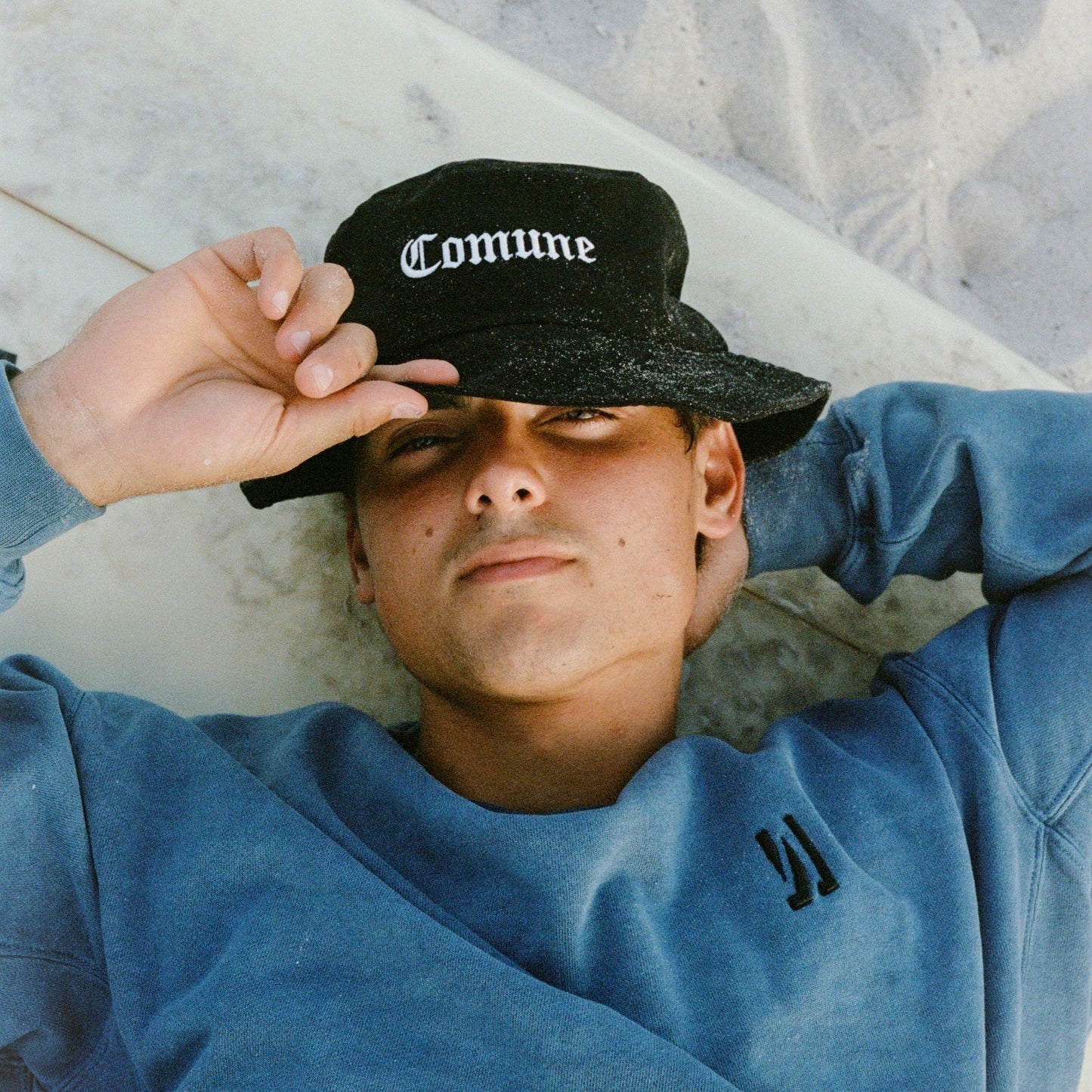 Person wearing a black 'Comune' hat and Comune's blue sweatshirt lying at the beach sand.