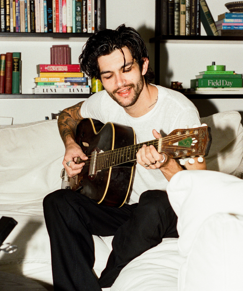 COMUNE artist Brandon Shoop playing a guitar in a cozy room with bookshelves in the background