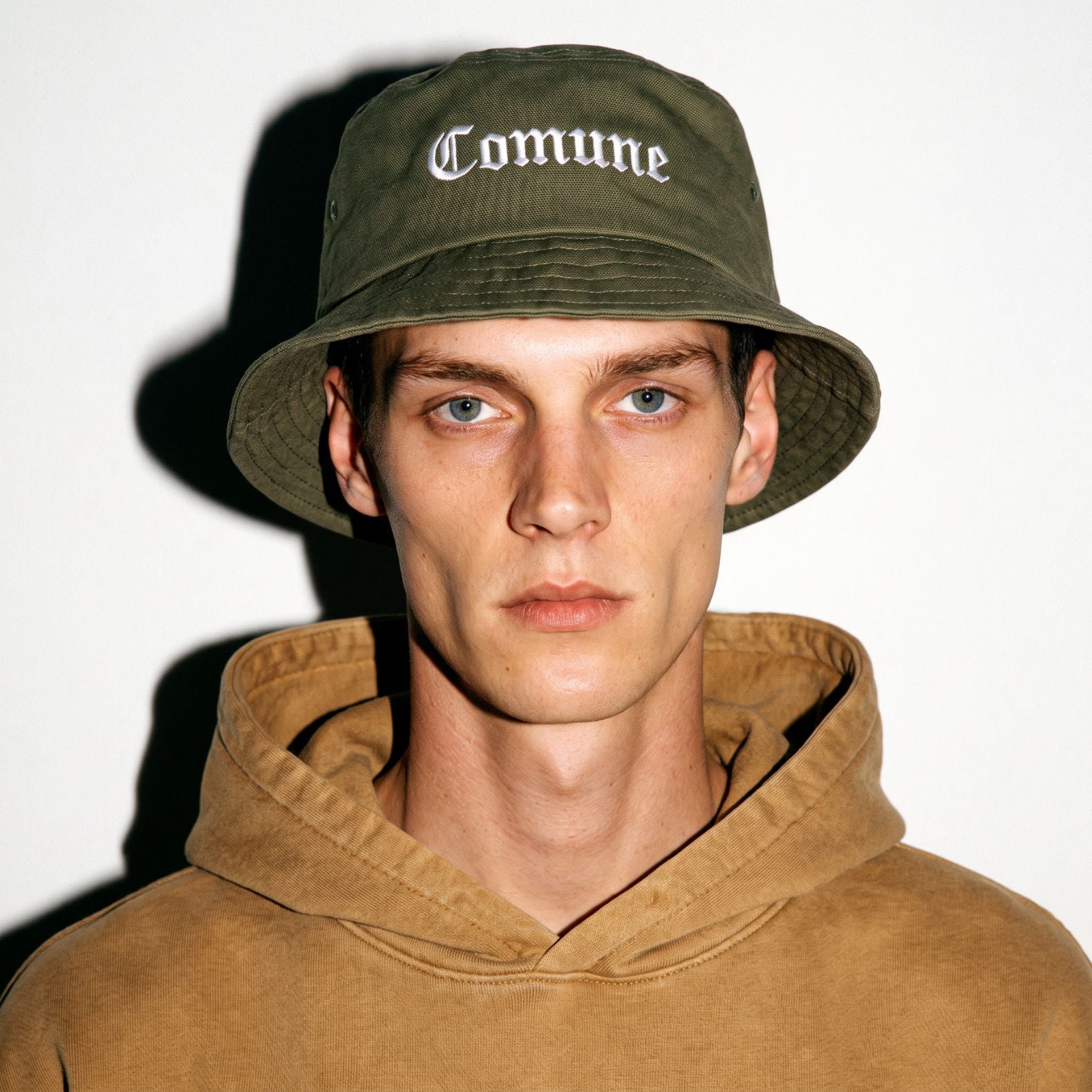 A man wearing a army green COMUNE Cali bucket hat with 'Comune' embroidered in white Gothic lettering, paired with a brown hooded sweatshirt, set against a plain white background with a strong shadow.