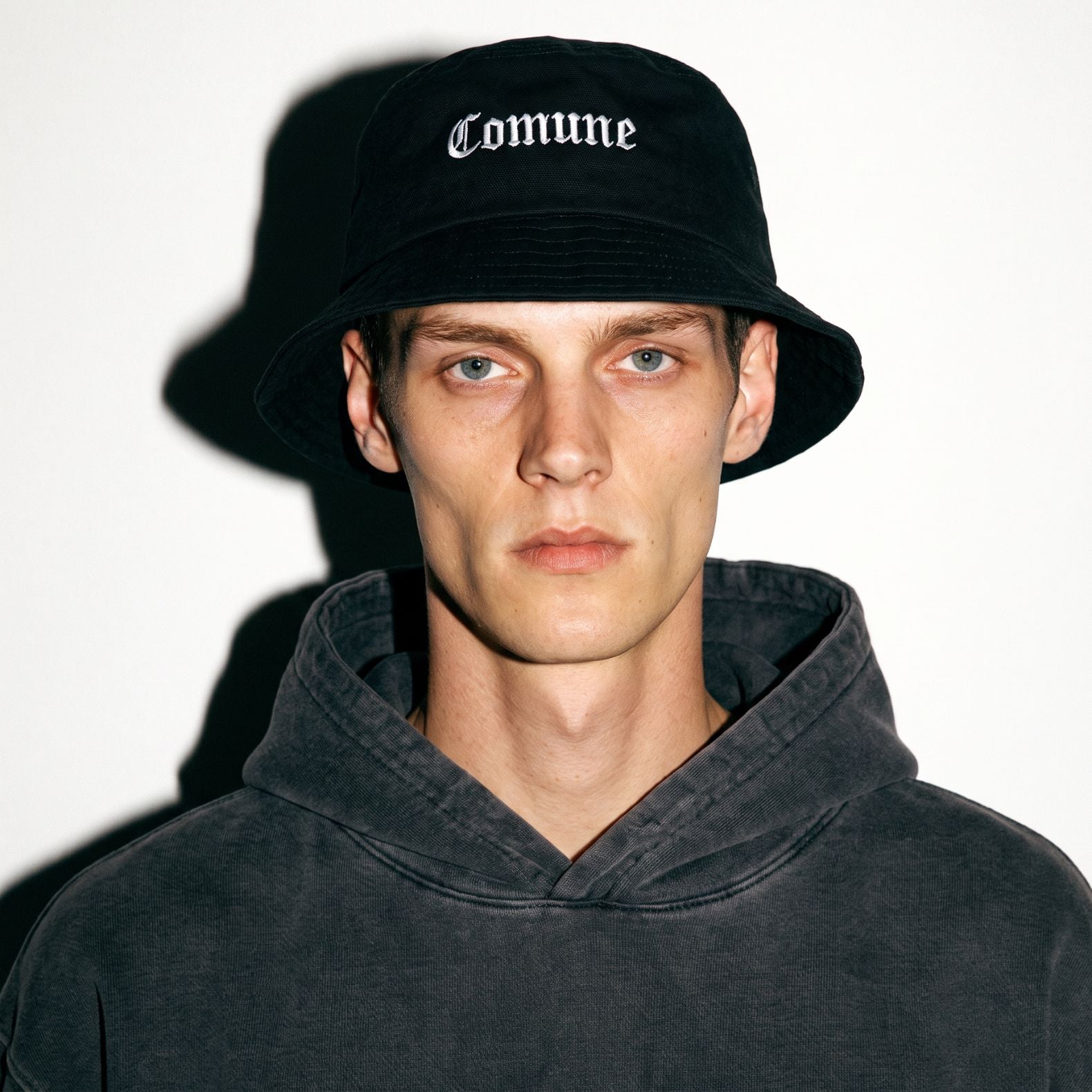 A man wearing a black COMUNE Cali bucket hat with 'Comune' embroidered in white Gothic lettering and a dark gray washed hoodie, photographed against a white background with a strong shadow.