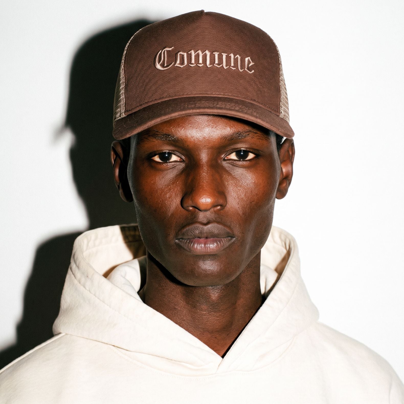 A close-up portrait of a model wearing a brown COMUNE Cali trucker-style cap with 'Comune' embroidered in a gothic font on the front, paired with a cream-colored hoodie against a white background with a strong shadow.