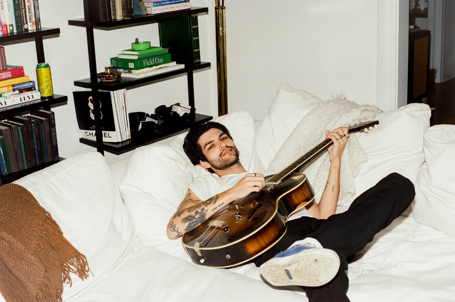 Man lying on a bed holding a guitar in a cozy room with bookshelves.
