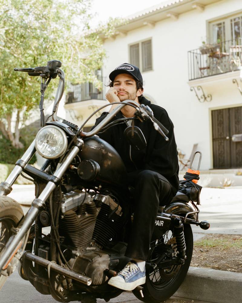 COMUNE artist Brandon Shoop sitting on a motorcycle in front of a house