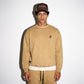 A man wearing a acorn color COMUNE Duke Crew sweatshirt with a small black embroidered logo on the left chest, matching tan sweatpants, and a brown baseball cap with 'Comune' embroidered on it, standing against a white background.