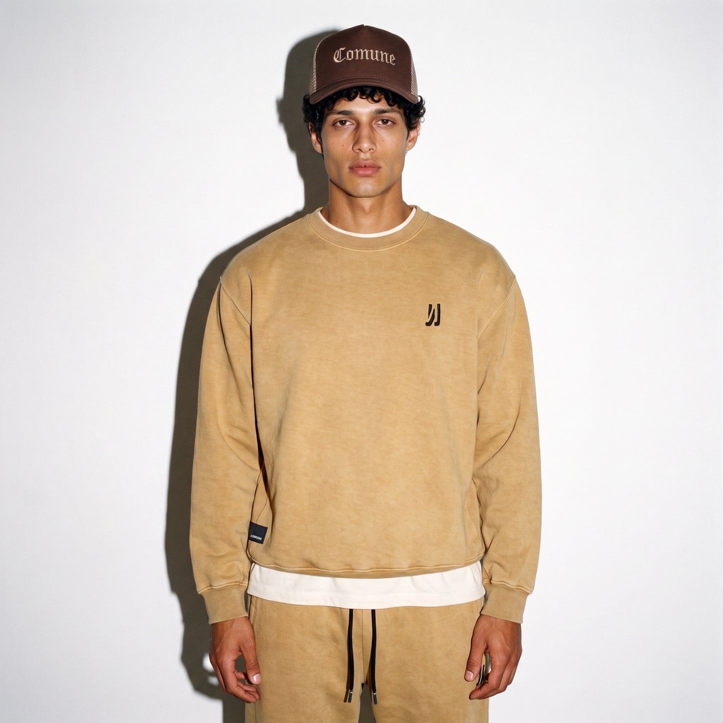 A man wearing a acorn color COMUNE Duke Crew sweatshirt with a small black embroidered logo on the left chest, matching tan sweatpants, and a brown baseball cap with 'Comune' embroidered on it, standing against a white background.