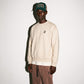 A model wearing a COMUNE Crew sweatshirt with a small black logo on the left chest in bone color, paired with brown drawstring sweatpants and a green trucker hat, standing against a plain off-white wall.