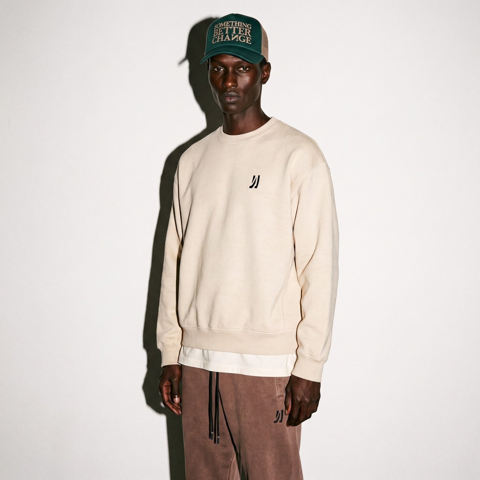 A model wearing a COMUNE Crew sweatshirt with a small black logo on the left chest in bone color, paired with brown drawstring sweatpants and a green trucker hat, standing against a plain off-white wall.