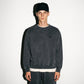 A man wearing a faded black washed COMUNE Duke Crew sweatshirt, matching sweatpants, a black baseball cap with tonal black 'COMUNE' embroidered, and a white undershirt, standing against a plain white wall.