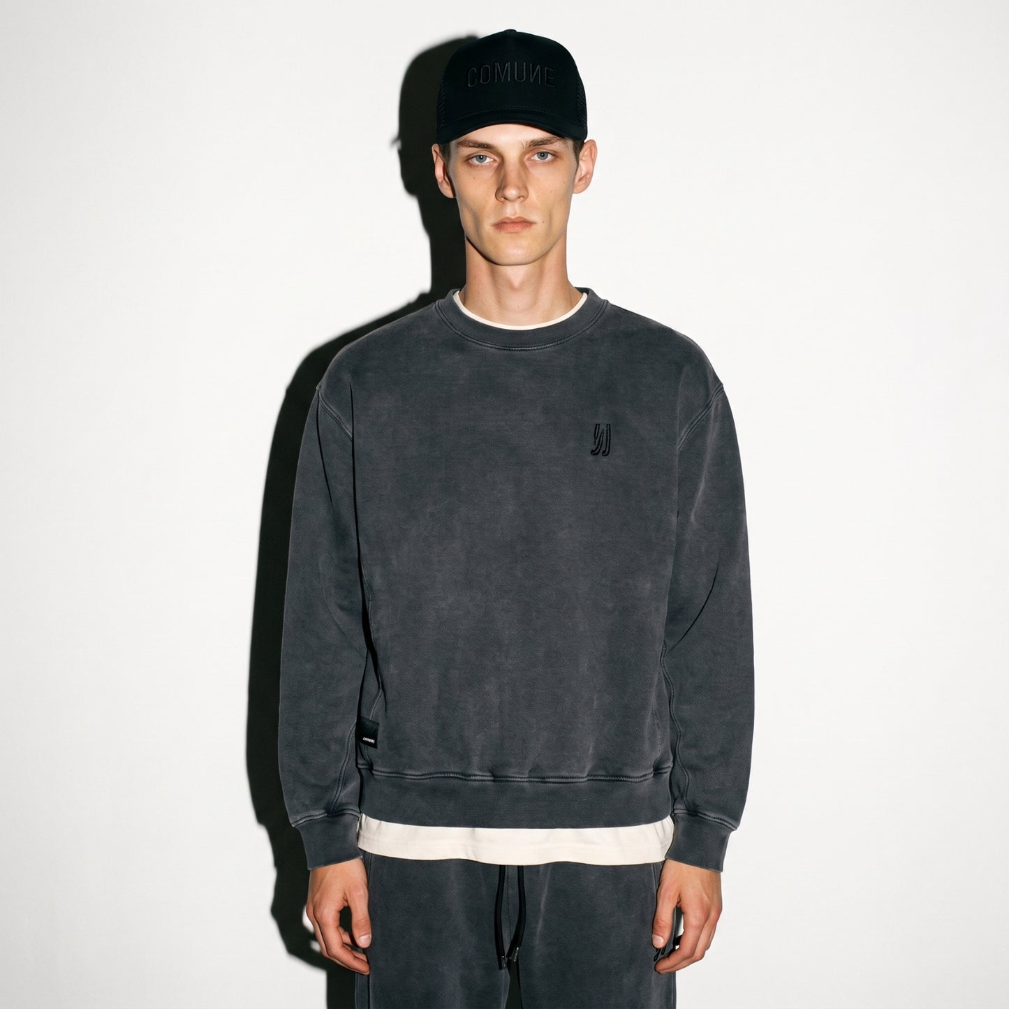 A man wearing a faded black washed COMUNE Duke Crew sweatshirt, matching sweatpants, a black baseball cap with tonal black 'COMUNE' embroidered, and a white undershirt, standing against a plain white wall.