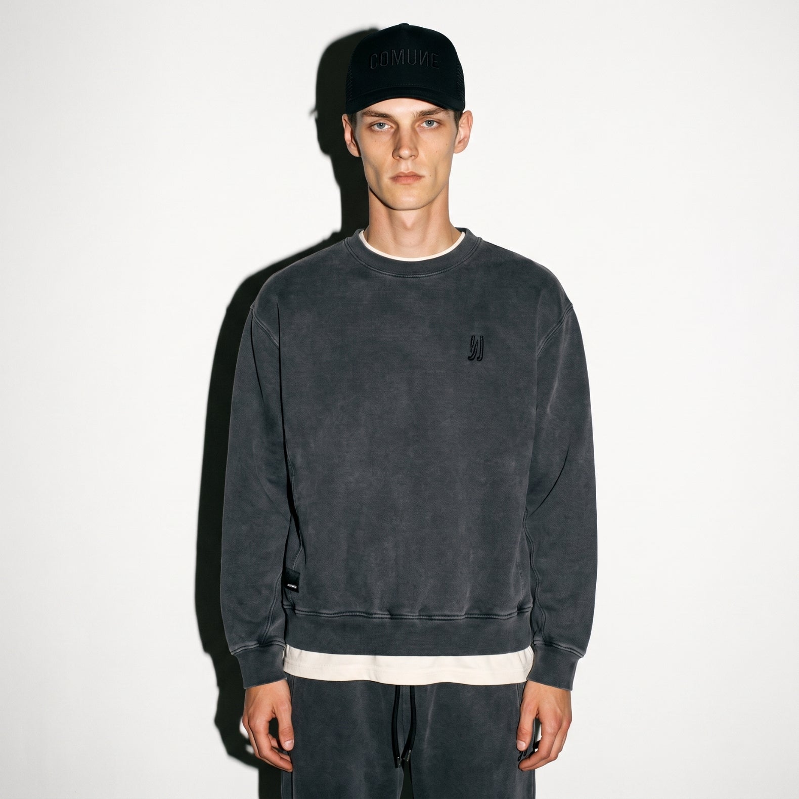 A man wearing a faded black washed COMUNE Duke Crew sweatshirt, matching sweatpants, a black baseball cap with tonal black 'COMUNE' embroidered, and a white undershirt, standing against a plain white wall.
