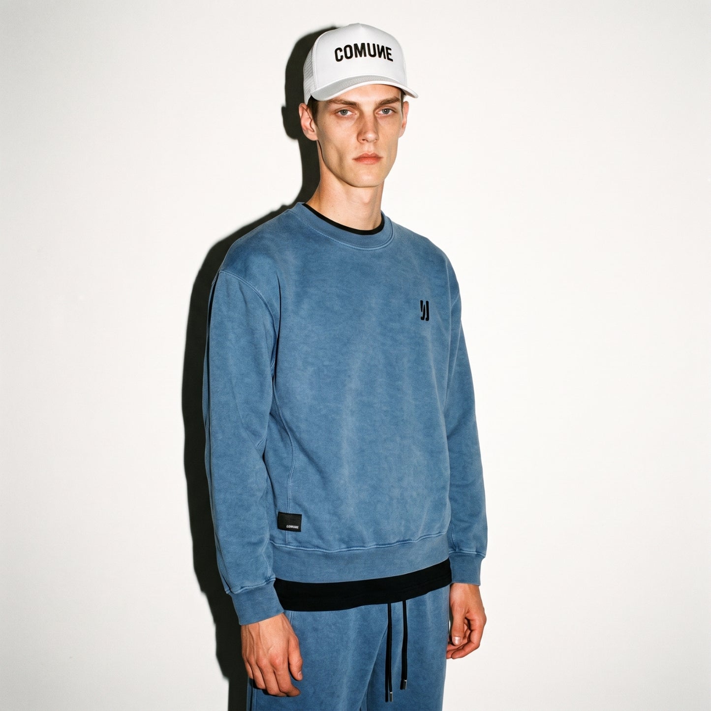A man wearing a marine washed COMUNE Duke Crew sweatshirt and matching sweatpants with black trim, paired with a white cap that says COMUNE, standing against a plain white wall.