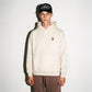A model wearing a COMUNE Duke hoodie in color bone with a small black embroidered logo on the left chest, paired with brown pants and a black trucker hat with "COMUNE" patch on it, standing against a plain white wall.