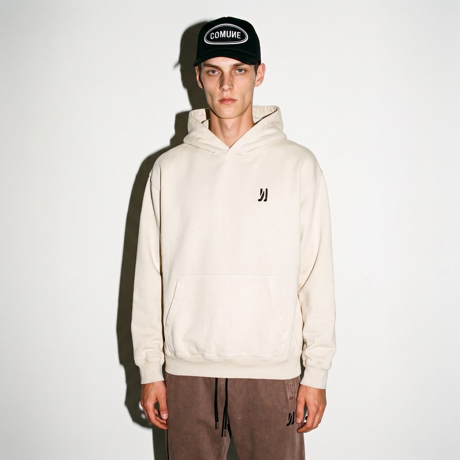 A model wearing a COMUNE Duke hoodie in color bone with a small black embroidered logo on the left chest, paired with brown pants and a black trucker hat with "COMUNE" patch on it, standing against a plain white wall.
