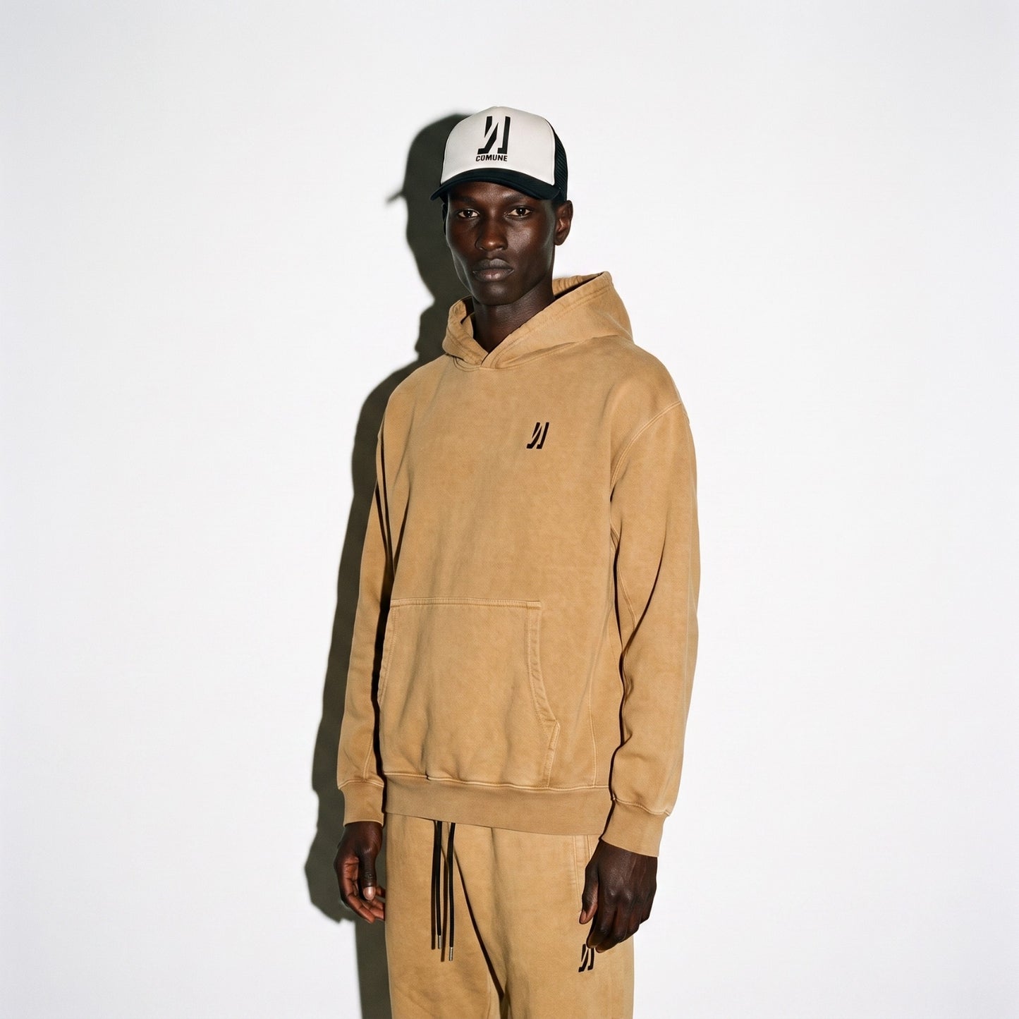A man wearing COMUNE Duke hoodie in camel brown color and matching sweatpants set with a black embroidered logo, paired with a black and white Icon COMUNE logo trucker hat, against a plain white background.