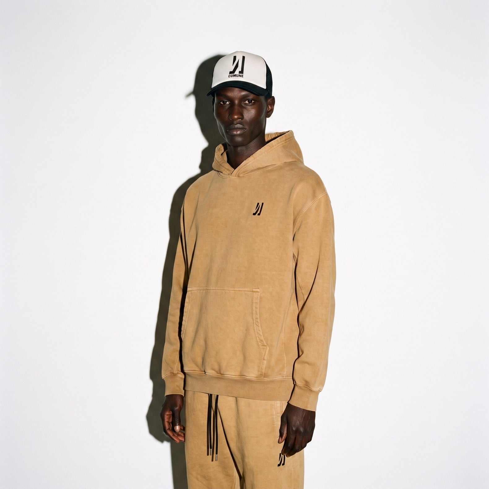 A man wearing COMUNE Duke hoodie in camel brown color and matching sweatpants set with a black embroidered logo, paired with a black and white Icon COMUNE logo trucker hat, against a plain white background.