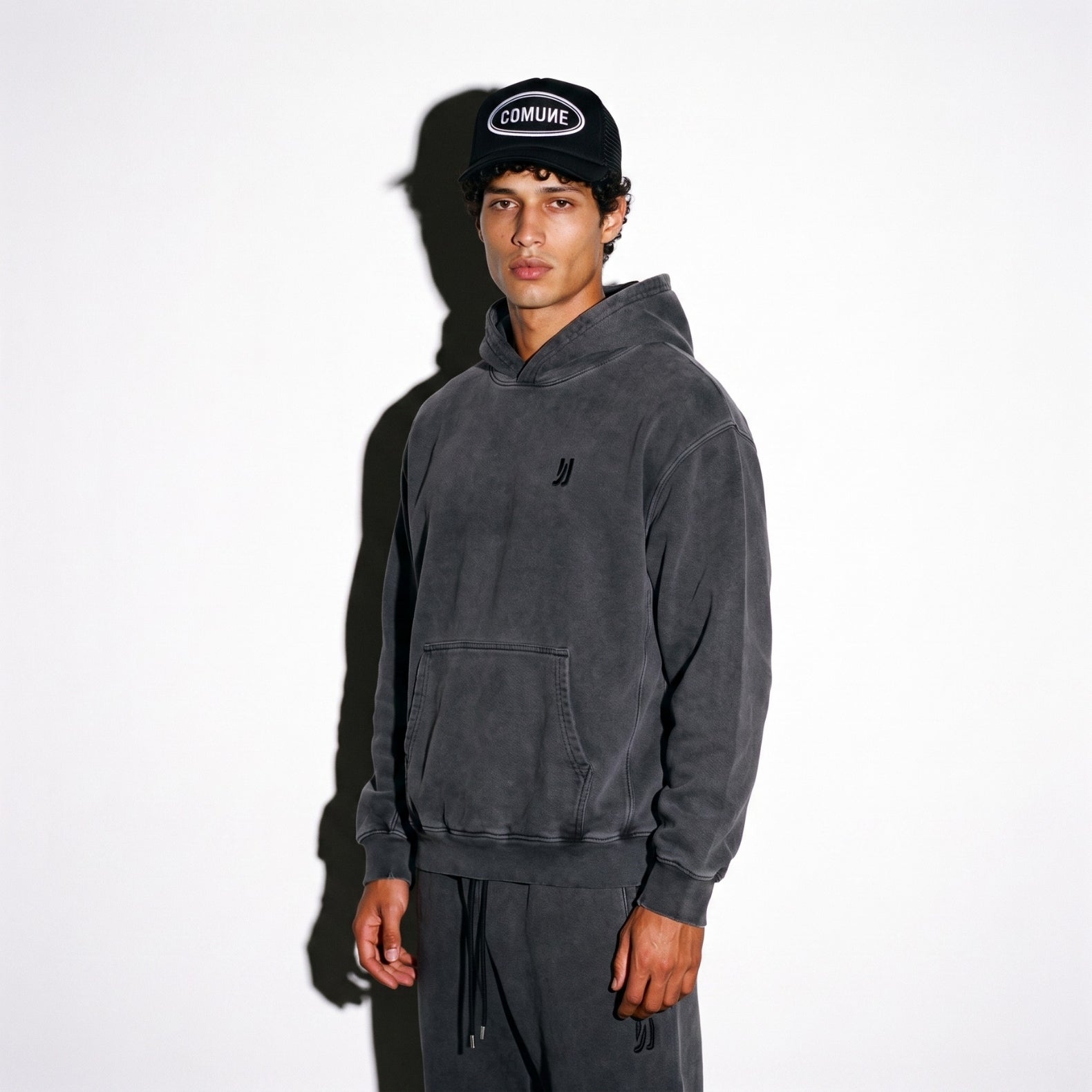 A man wearing a COMUNE faded black washed hoodie with a small black logo on the chest, matching dark sweatpants, and a black trucker hat with 'COMUNE' patch on it, standing against a white background with a strong shadow.
