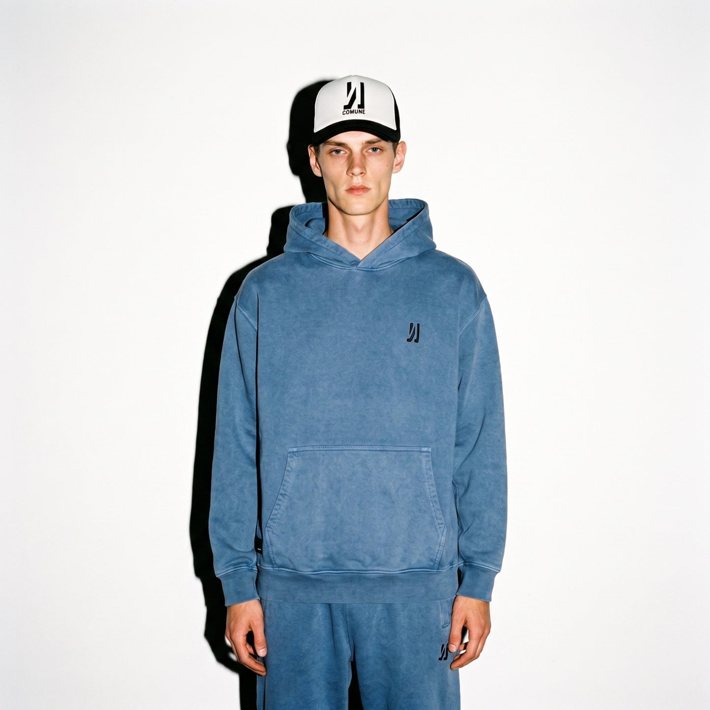 A man wearing a marine COMUNE hooded sweatshirt and matching sweatpants with a black and white Icon Trucker Hat, standing against a plain white background.