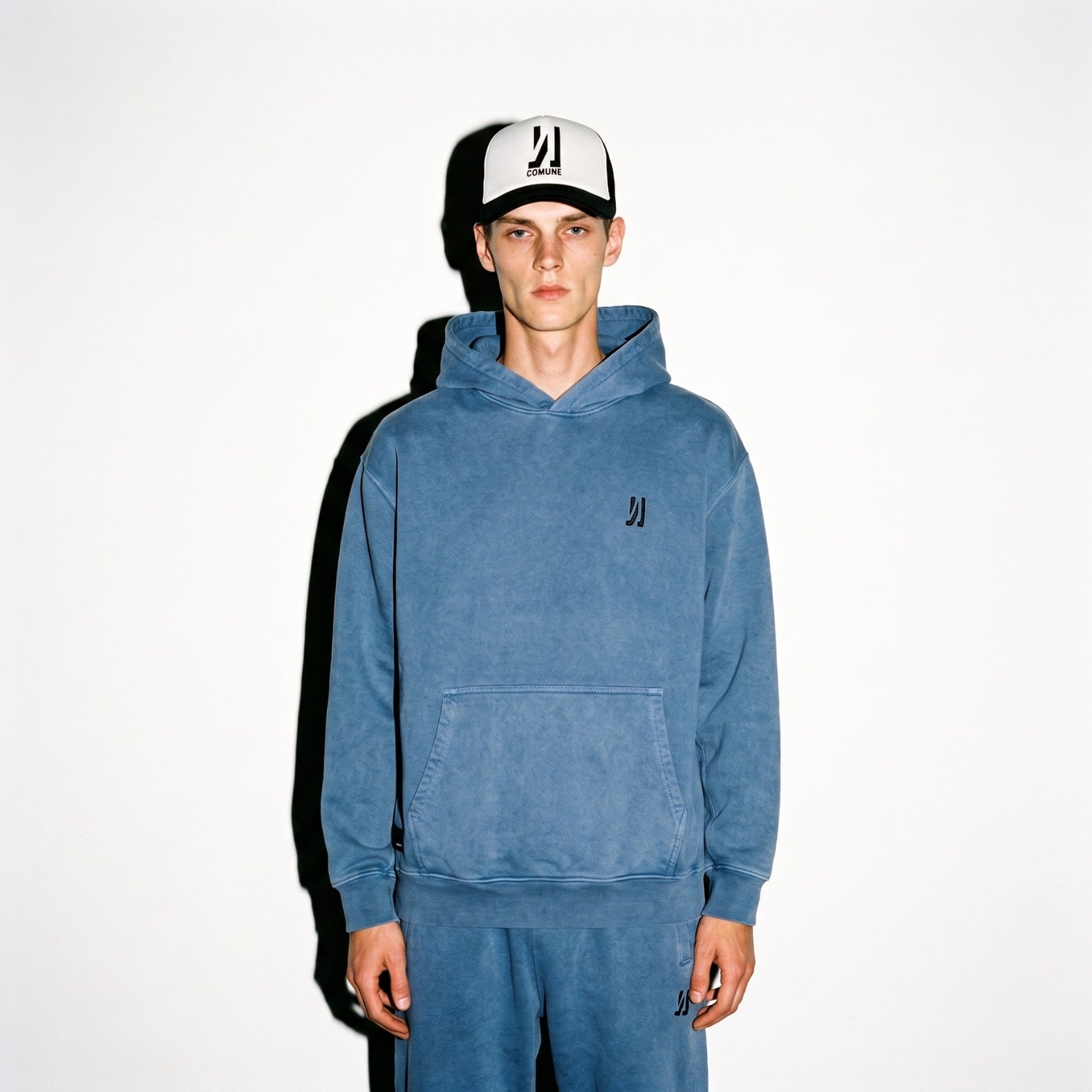A man wearing a marine COMUNE hooded sweatshirt and matching sweatpants with a black and white Icon Trucker Hat, standing against a plain white background.
