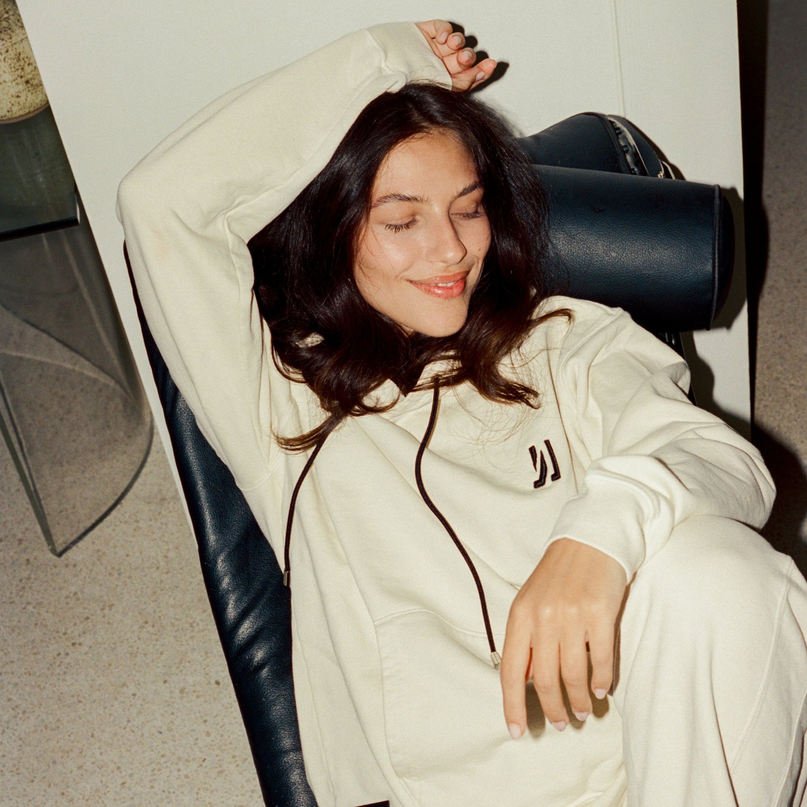 Person wearing a 'COMUNE' white hoodie sitting on a black chair indoors.