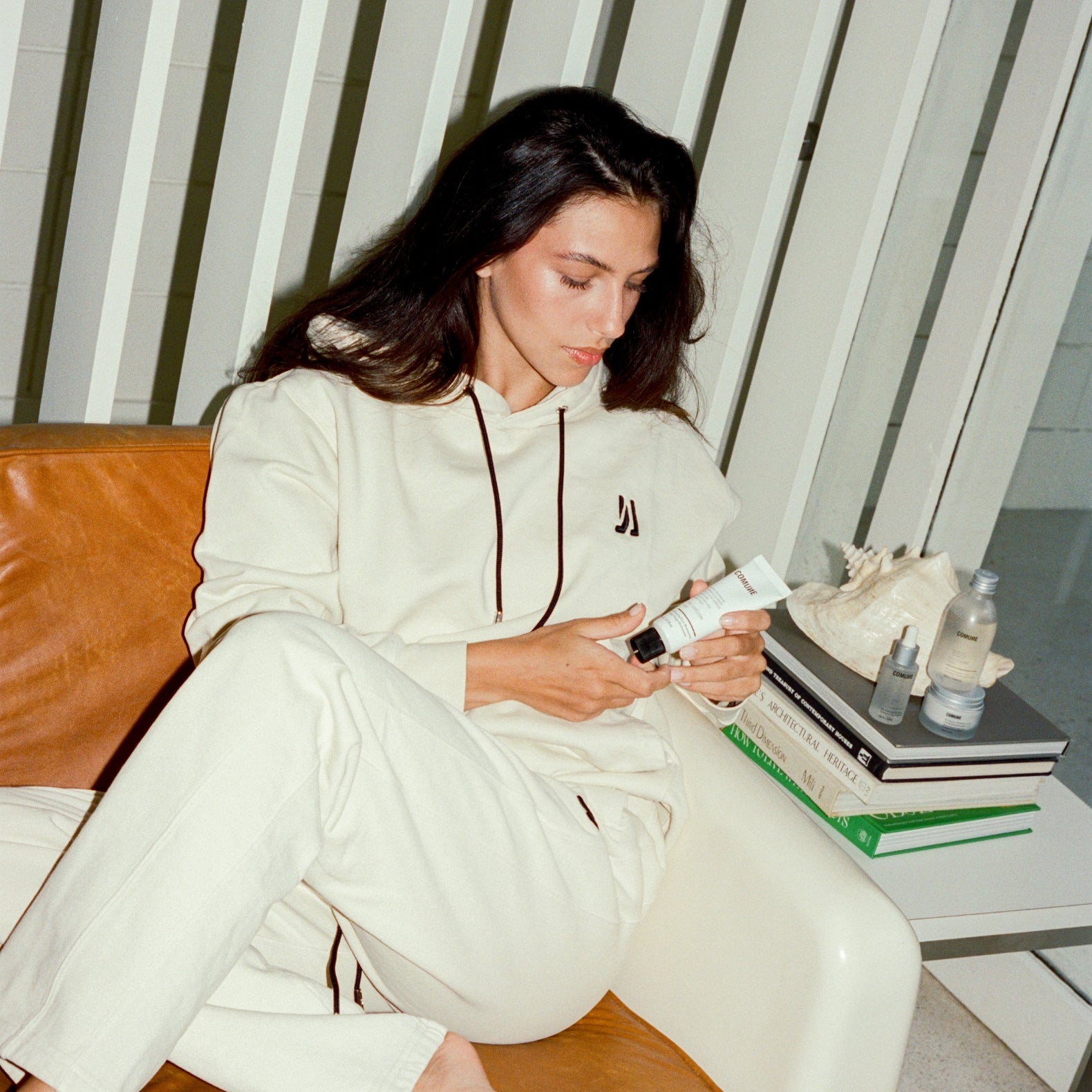 Woman in a white 'COMUNE' tracksuit sitting on a couch holding comune hydra shroom cream with a striped wall in the background.