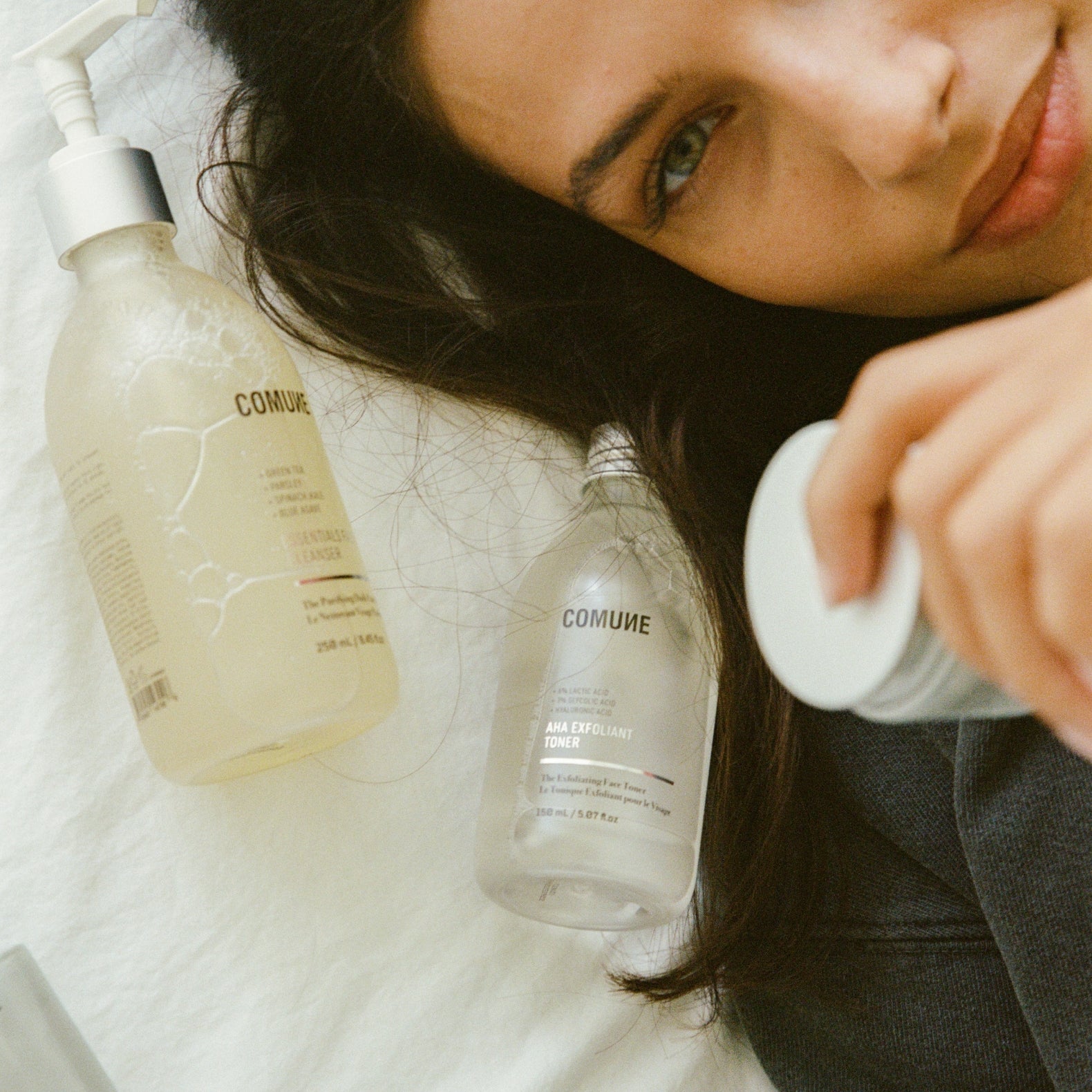 Woman holding a bottle of 'COMUNE' product with another bottle on the bed