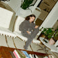 Person sitting on a white chair wearing comune gear in a room with plants and furniture, holding 'COMUNE' Essentials Skincare products.  