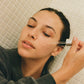 Person applying a 'COMUNE' dropper of liquid to their face in a bathroom setting.