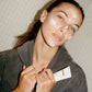 Woman wearing a COMUNE hoodie holding a product label 'COMUNE' hydra shroom cream, against a tiled wall background.