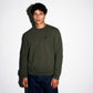 A man wearing an army green COMUNE Luka Crew sweatshirt with a black embroidered logo on the left chest, paired with dark blue jeans, standing against a white wall with a soft shadow.