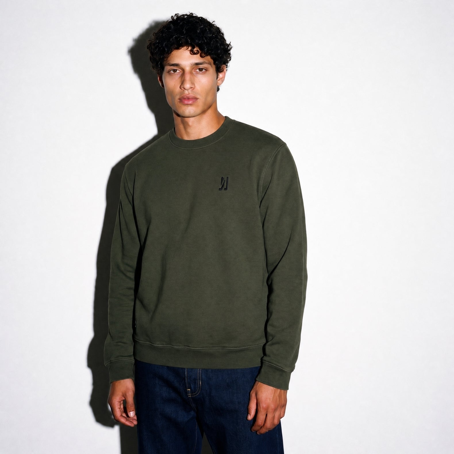 A man wearing an army green COMUNE Luka Crew sweatshirt with a black embroidered logo on the left chest, paired with dark blue jeans, standing against a white wall with a soft shadow.
