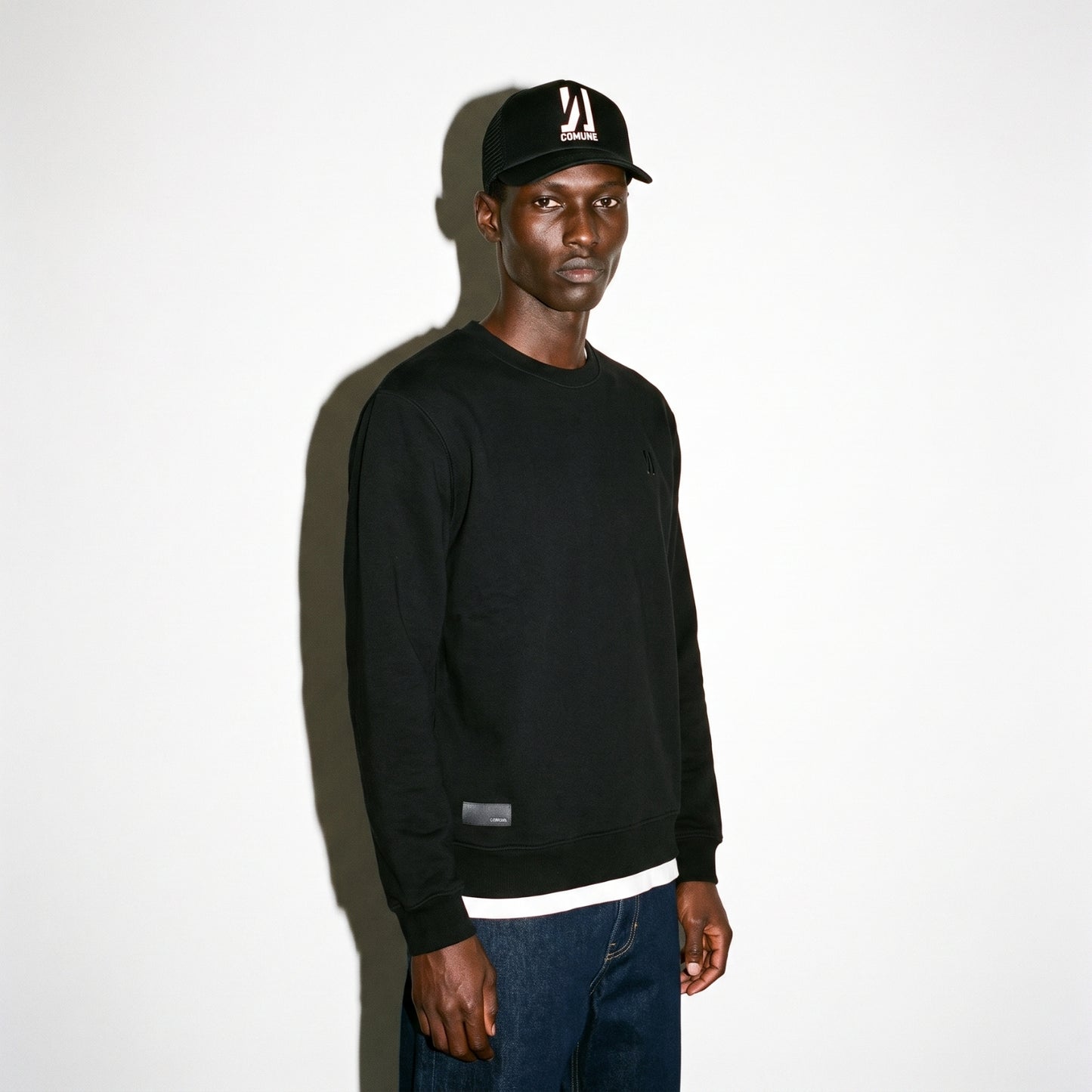A man wearing a black COMUNE Luka Crew sweatshirt, dark blue jeans, and a black baseball cap with a white graphic logo, standing against a plain white wall with a strong shadow.