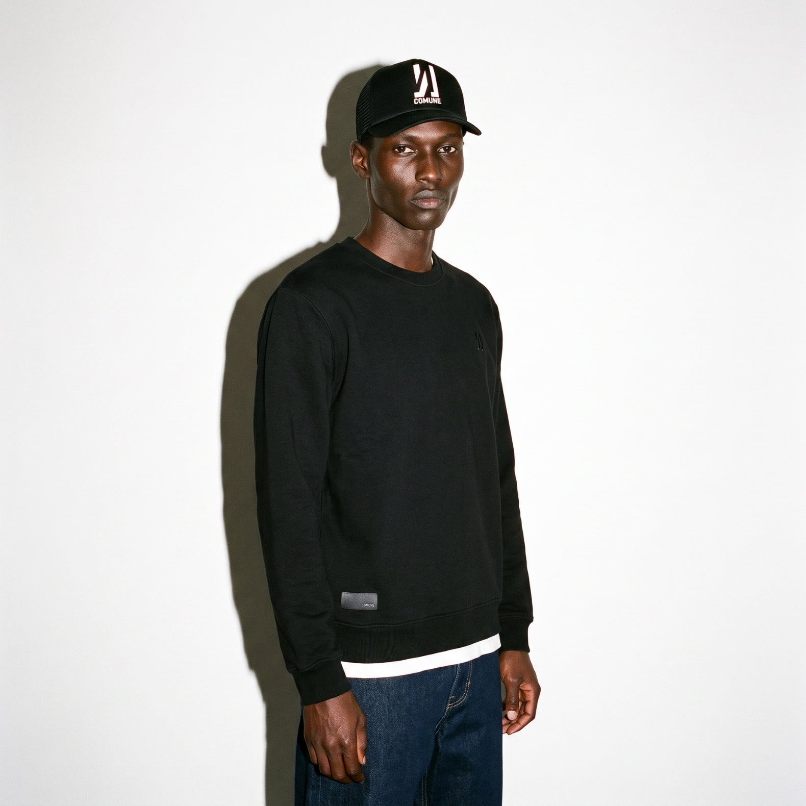 A man wearing a black COMUNE Luka Crew sweatshirt, dark blue jeans, and a black baseball cap with a white graphic logo, standing against a plain white wall with a strong shadow.