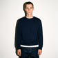 A man wearing a dark navy COMUNE Luka Crew sweatshirt with a embroidered logo on the left chest, layered over a white t-shirt and dark blue jeans, standing against a plain white wall.