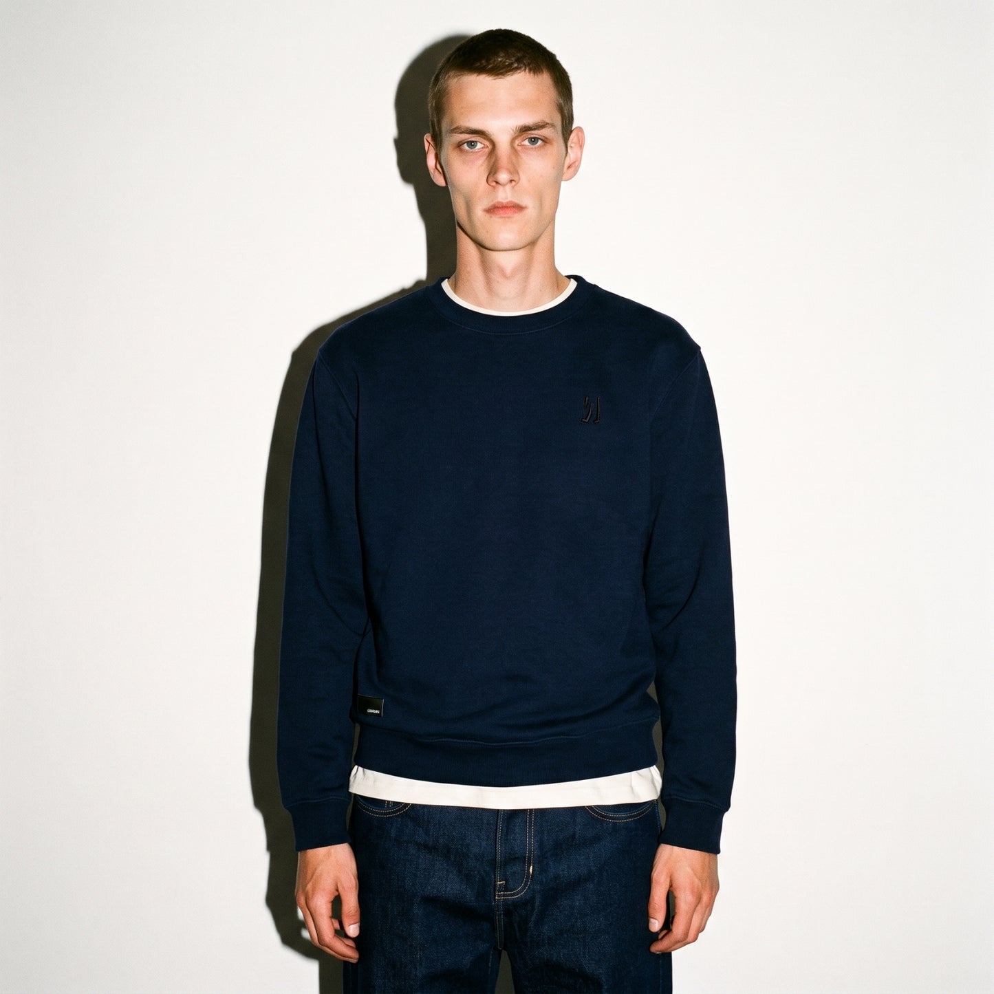 A man wearing a dark navy COMUNE Luka Crew sweatshirt with a embroidered logo on the left chest, layered over a white t-shirt and dark blue jeans, standing against a plain white wall.