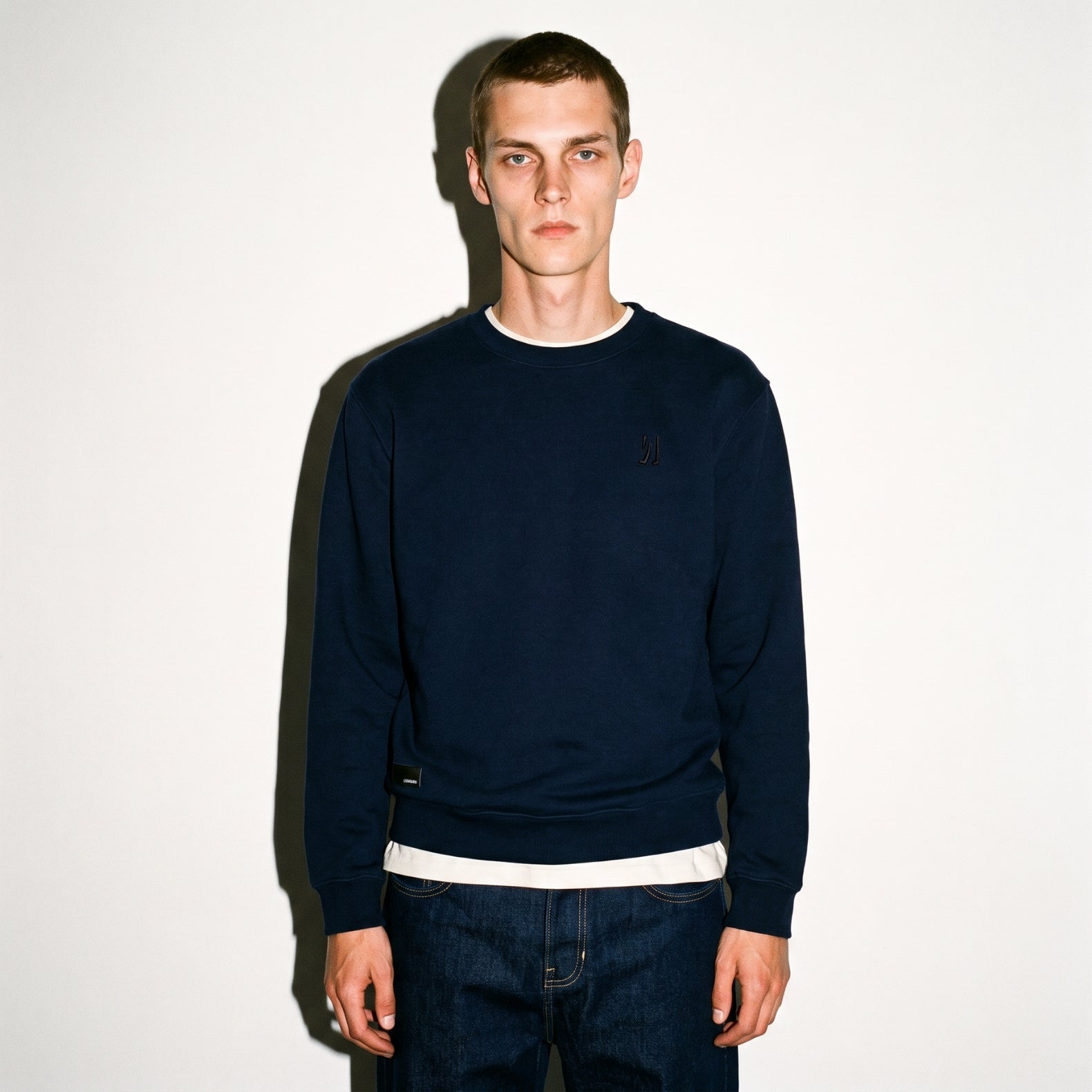 A man wearing a dark navy COMUNE Luka Crew sweatshirt with a embroidered logo on the left chest, layered over a white t-shirt and dark blue jeans, standing against a plain white wall.