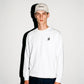 A man wearing a white COMUNE Luka Crew sweatshirt with a small black logo on the left chest, paired with a beige baseball cap and dark jeans, standing against a white wall with a hard shadow.