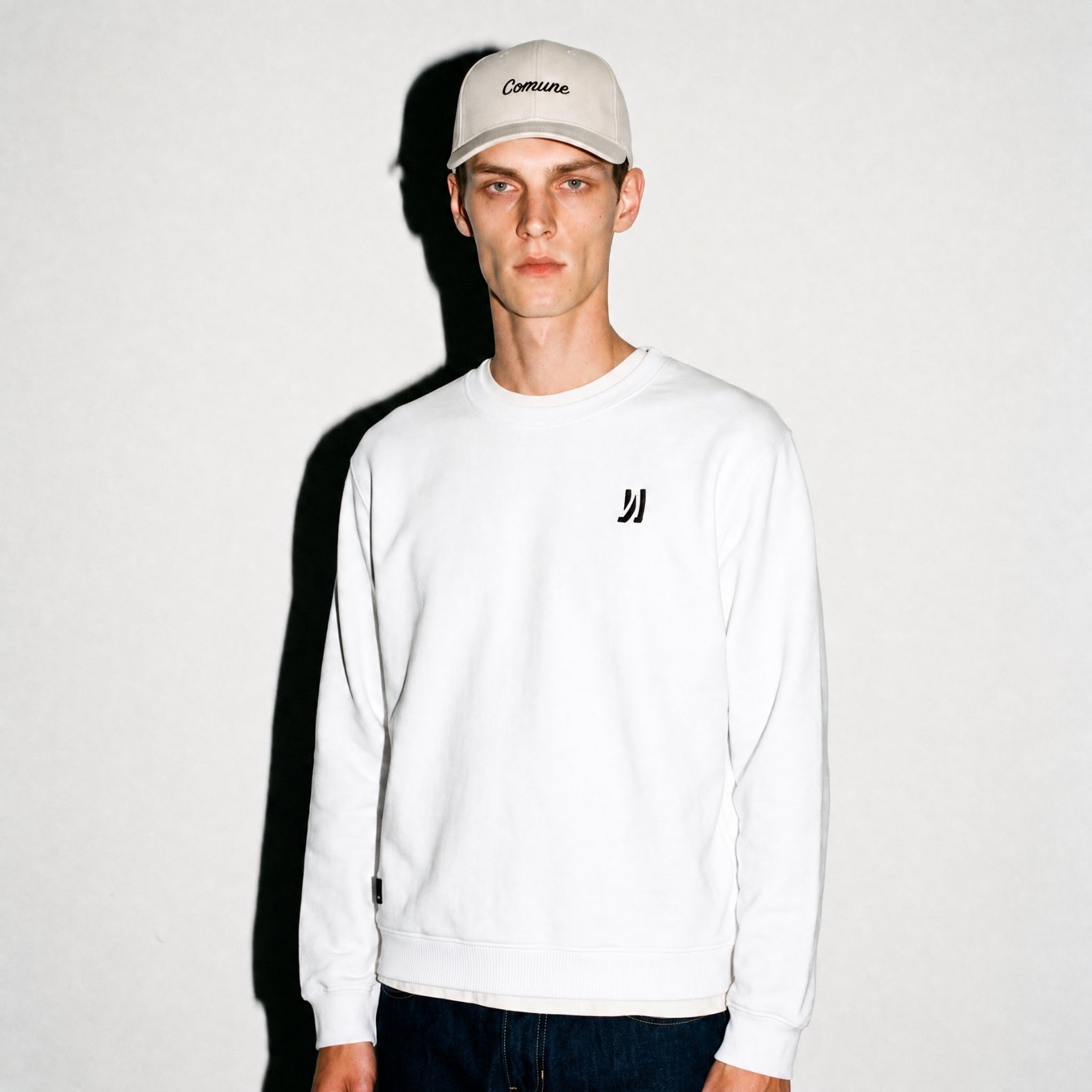 A man wearing a white COMUNE Luka Crew sweatshirt with a small black logo on the left chest, paired with a beige baseball cap and dark jeans, standing against a white wall with a hard shadow.