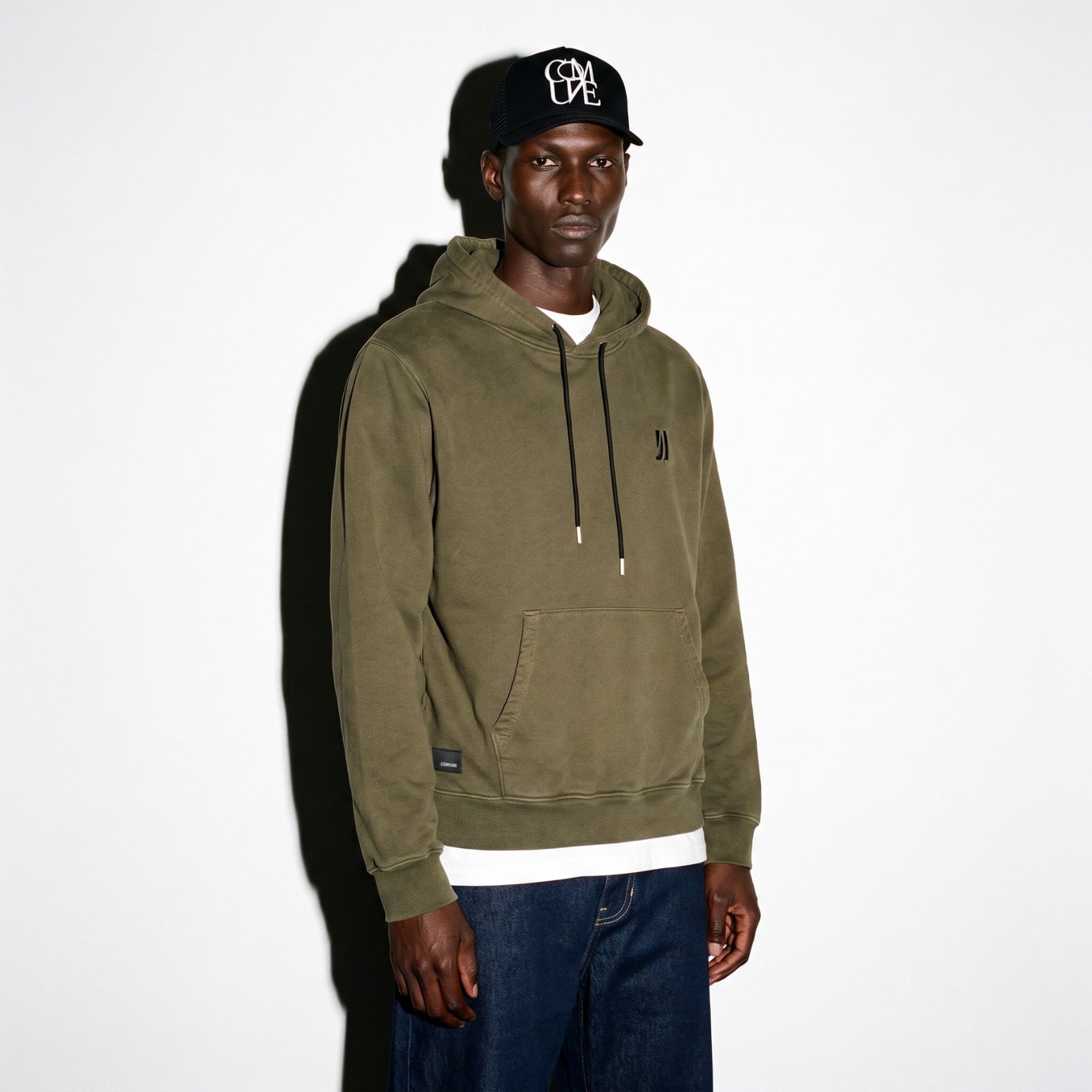 A man wearing an olive green COMUNE Luka pullover hoodie with a small black embroidered logo on the left chest, paired with a black baseball cap and dark blue jeans, standing against a white wall.